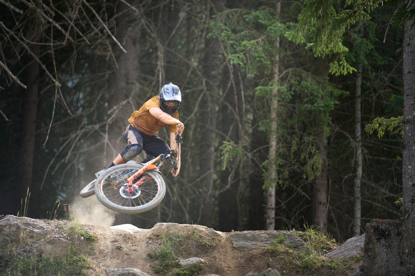 A mountain biker in a helmet and orange shirt jumps a bike over a rocky forest trail